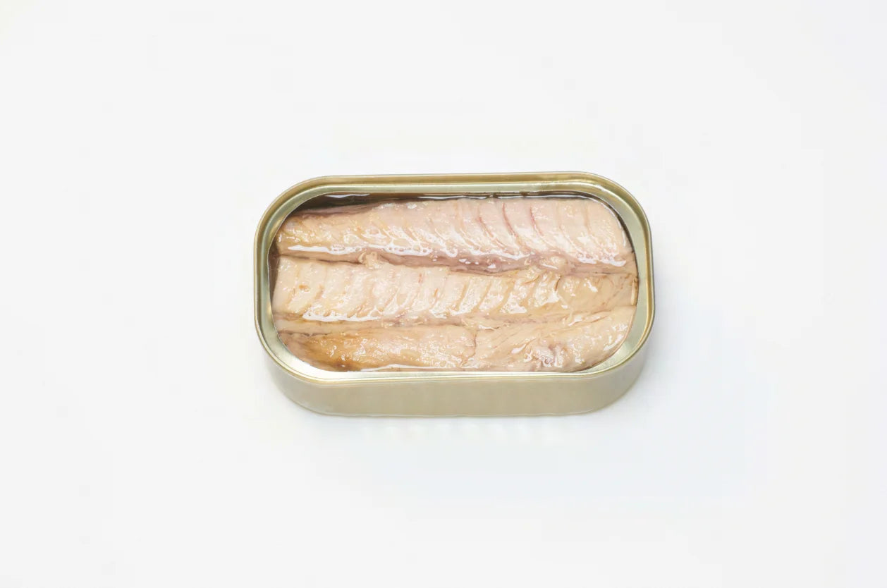 A photorealistic close-up of a white tray containing three PYSCIS cans labeled "3-Pack Little Bullet Tuna Filets" (SKU: AUX22). The label features the PYSCIS brand logo and highlights the product's use of sustainable fishing practices. A blue and green gradient adorns the label in the background.  The image focuses on a single can tilted slightly forward, revealing a glimpse of the flaky, white tuna meat bathed in olive oil through the transparent window in the can.