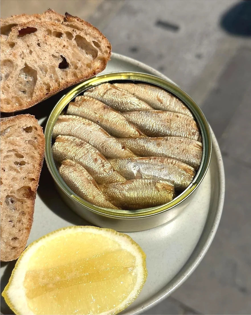 An opened tin of PYSCIS sprats in olive oil from Spain. The tin is made of natural paper and rubber and has a handmade appearance. The tin contains the top ten sprats swimming in olive oil. The olive oil is clear and has a golden color.